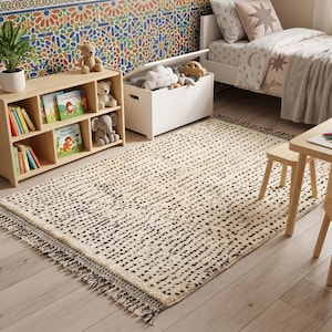 May include: A cream-colored rug with a dotted black pattern and fringed edges. The rug is in a child's bedroom, alongside a wooden bookshelf with books, a white toy chest, and a small wooden table with crayons.