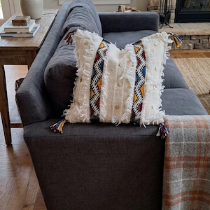 Peut inclure: Un coussin carré en tissu texturé couleur crème, orné de bandes verticales aux motifs géométriques rouges, jaunes et bleus. Le coussin est agrémenté de bords effilochés, de petits boutons en bois ronds et de pompons à chaque coin.