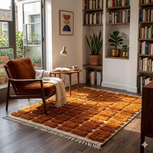 May include: An orange and cream-colored rug with a geometric pattern. The rug is rectangular with a fringe border. A brown armchair and a small wooden table with a lamp and books are in the background. A cozy living room setting.