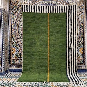 May include: A green rectangular rug with a central yellow stripe and a black and white striped border. The rug has a textured surface and a fringe along the bottom edge. The rug is displayed against a backdrop of ornate tilework.