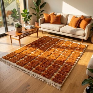 May include: An orange and cream-colored area rug with a geometric pattern of rectangular blocks. The rug has a fringed edge and is placed on a hardwood floor. The rug is in a living room setting with a sofa and pillows.
