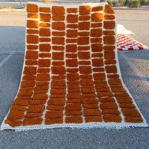 May include: A rectangular, handmade rug with a repeating pattern of rust-colored, rectangular tufts against a white background. The rug is made of a thick, textured material and has a fringed edge. The rug is displayed outdoors on a concrete surface.
