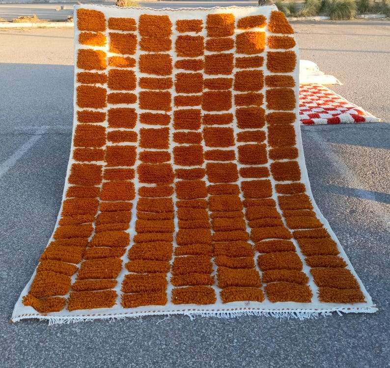 May include: A rectangular, handmade rug with a repeating pattern of rust-colored, rectangular tufts against a white background. The rug is made of a thick, textured material and has a fringed edge. The rug is displayed outdoors on a concrete surface.