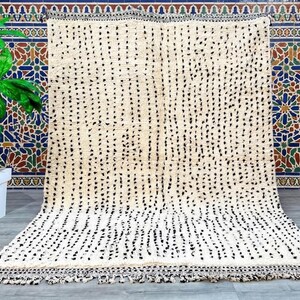 May include: A large, off-white rug with a pattern of black dots arranged in vertical lines. The rug has a fringe along the bottom edge and is displayed against a colorful tile background. The rug is a decorative floor covering.