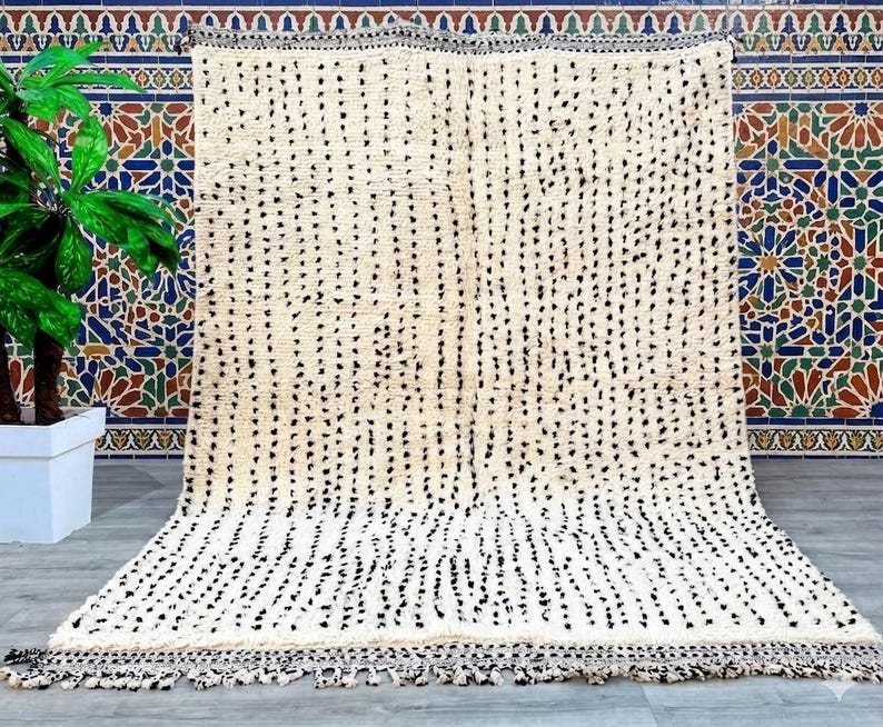 May include: A large, off-white rug with a pattern of black dots arranged in vertical lines. The rug has a fringe along the bottom edge and is displayed against a colorful tile background. The rug is a decorative floor covering.
