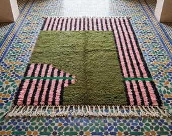 Peut inclure: Un tapis rectangulaire présentant un grand bloc central vert olive contrasté par des sections de rayures verticales roses et noires. Le tapis a un poil épais et texturé et est fini avec une longue frange de couleur crème sur le bord inférieur.