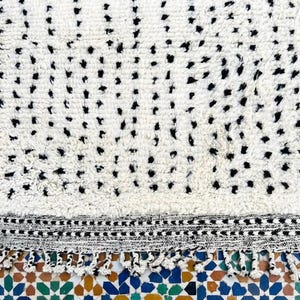 May include: A white rug with a pattern of black dots. The rug has a textured surface and a black and white border with fringe. The rug is placed on a colorful tile surface.