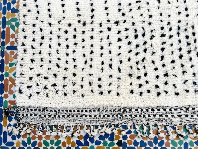 May include: A white rug with a pattern of black dots. The rug has a textured surface and a black and white border with fringe. The rug is placed on a colorful tile surface.