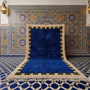 May include: A rectangular area rug featuring a large, solid deep blue center panel. The rug is framed by a border of alternating olive green squares and a wide, light beige outer edge. The short ends of the rug are finished with white fringe.