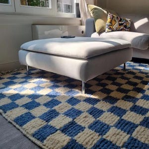 Könnte beinhalten: Ein hellgrauer Hocker mit Chrombeinen steht auf einem blau-cremefarbenen Schachbrettmuster-Teppich. Im Hintergrund befindet sich ein graues Sofa mit Kissen. Der Raum ist gut beleuchtet.