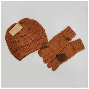 May include: A burnt orange knit beanie and a pair of matching knit gloves. The beanie features a ribbed texture and a tan tag with the text C.C. The gloves have a ribbed cuff, a brown patch on the palm, and a small brown tag with the text C.C.