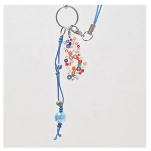 May include: A clear, abstract-shaped keychain charm with colorful circular and square accents. The charm is attached to a blue cord with decorative beads and a silver-tone clasp. The keychain is designed to hold keys or be used as a decorative accessory.