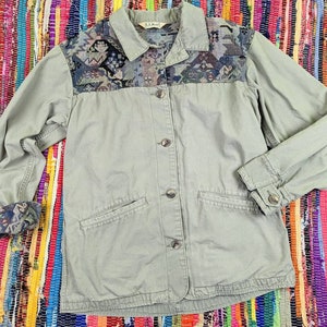 May include: A green utility jacket with a patterned fabric on the shoulders and a button closure. The jacket has two pockets on the front and a single pocket on the left sleeve. The label on the jacket reads "L.L.Bean".