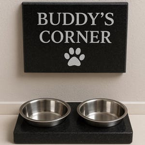 Peut inclure: Un présentoir noir pour animaux de compagnie avec deux bols en acier inoxydable. Le panneau au-dessus indique "BUDDY'S CORNER" avec une empreinte de patte. Les bols reposent sur une base noire assortie.