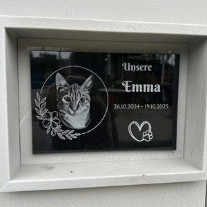 May include: A rectangular black memorial plaque set in a light grey stone frame. The plaque features an engraved portrait of a cat inside a circular border with floral accents, the text "Unsere Emma", the dates "26.10.2024 - 19.10.2025", and a heart icon.