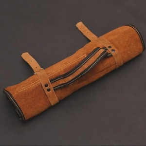 Op de afbeelding: Een bruine leren gereedschapsrol met twee zwarte strepen en witte stiksels. De rol wordt vastgezet met twee leren riemen en gespen. De gereedschapsrol is ontworpen om gereedschap in op te bergen.