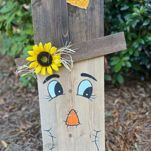 May include: A wooden scarecrow face with painted eyes, nose, and mouth. The scarecrow has a sunflower and straw hat on top.