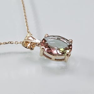 May include: A gold necklace with an oval gemstone pendant. The gemstone has a gradient of pink, green, and grey hues. The pendant is held in a gold setting and suspended from a fine chain. The necklace is a piece of jewellery.