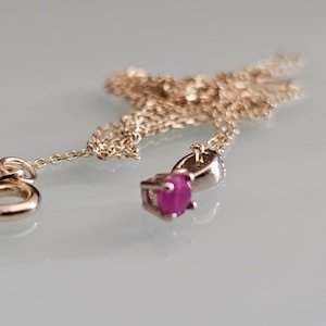 Puede incluir: Un collar de color dorado con un pequeño colgante con una vibrante gema rosa de corte ovalado. La cadena es delicada y el colgante está engastado en un metal plateado. El collar se exhibe sobre una superficie clara.