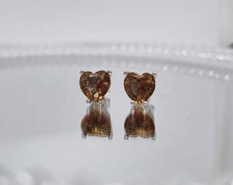 May include: A pair of stud earrings featuring heart-shaped, amber-colored gemstones. The gemstones are set in a silver-colored metal. The earrings are displayed on a reflective surface, creating a mirrored effect. The earrings are a jewelry item.