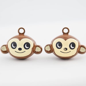 May include: Two brown monkey-shaped charms with a light beige face, black eyes, and a smiling mouth. Each charm has a small loop at the top for hanging. The charms are likely for jewelry or crafts.