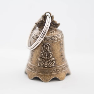 May include: A small, gold-colored bell with a silver ring. The bell has intricate carvings and a figure of a person sitting in a meditative pose. The bell is a traditional Buddhist item.
