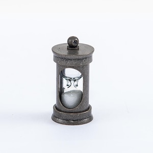 May include: A small, antique-style hourglass with a metal frame and a clear glass bulb. The hourglass is filled with white sand and is sitting on a white surface.