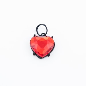 May include: A red heart-shaped charm with a black metal setting. The charm has a faceted design and is attached to a black metal ring.