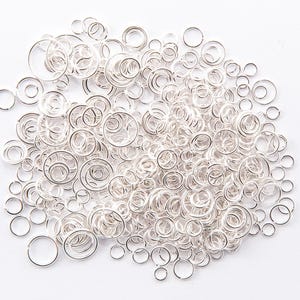 May include: A pile of small, shiny, silver jump rings of varying sizes, arranged on a white background. The rings are circular and likely made of metal, ideal for jewellery making or crafting projects.