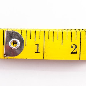 Può includere: Un nastro di misurazione giallo con un anello di metallo e segni neri. Il nastro indica "1" e "2".