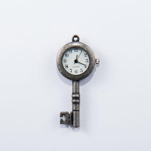 May include: A silver-coloured key pendant featuring a round clock face. The clock has black hands and numerals on a white background. The word "QUARTZ" is printed below the centre of the clock face. The key has a decorative handle and a keyhole design.