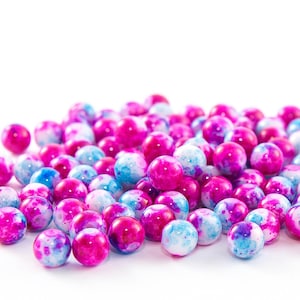 May include: A pile of round beads in shades of pink, blue, and white. The beads have a marbled appearance, with the colors blending together. The beads are scattered on a white surface, creating a visually appealing display.