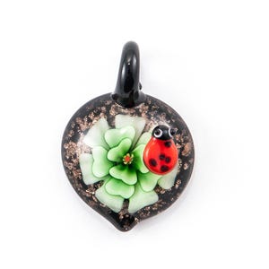 May include: A black glass pendant with a green flower and a red ladybug on top. The pendant has a gold glitter finish.