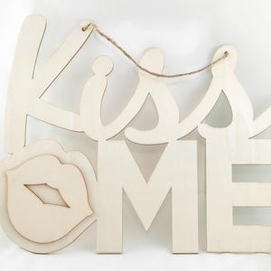 May include: Wooden sign with the words "Kiss Me" in a cursive font. The sign has a cutout of a pair of lips in the center.