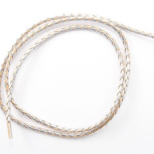 May include: Braided leather cord in a loop, featuring a mix of silver and tan strands. The cord is designed with a woven pattern, creating a textured appearance. The ends are finished with a smooth, solid color. Ideal for jewelry making or crafting.