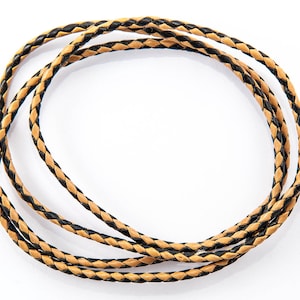 May include: Braided leather cord, arranged in a circular shape. The cord features a tan and black design, with the black sections forming a repeating pattern. This cord is likely used for jewellery making or crafts.