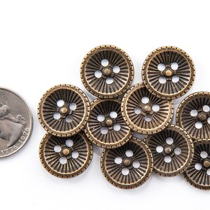 May include: A collection of antique brass-colored buttons with a radial design and four holes, next to a U.S. quarter coin. The buttons have a vintage aesthetic and are arranged on a white surface.