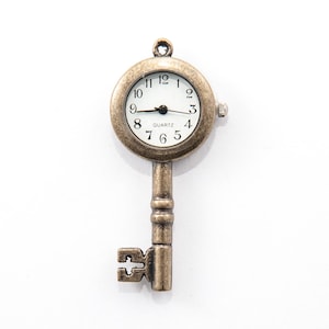 May include: A vintage-style key pendant with a clock face. The key is made of bronze-colored metal and features a round clock with black numerals on a white background. The clock hands point to approximately 8:40. The pendant is designed to be worn as jewelry.