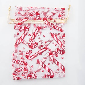 Puede incluir: Una bolsa de regalo de tul blanca con un patrón de bastón de caramelo rojo y blanco. La bolsa tiene un cordón de ajuste dorado.