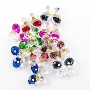 May include: A selection of clear glass butterfly charms in various colours. The charms are in shades of blue, green, red, pink, and brown, with gold glitter detailing. These decorative charms are suitable for jewellery making or craft projects.