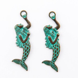 May include: Two bronze-colored mermaid charms with a verdigris finish. Each charm features a mermaid with flowing hair, a scaled tail, and an arm raised to hold a ring. The charms are ideal for jewelry making or crafting.