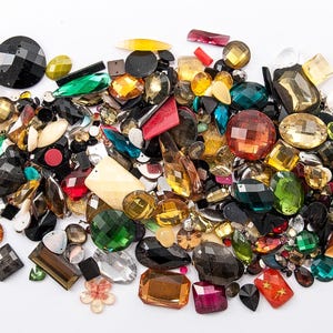 May include: A collection of colorful, faceted gemstones in various shapes and sizes. The assortment includes emerald green, ruby red, amber, and black stones, with some having a metallic sheen. The gems are scattered on a white surface, creating a vibrant display.