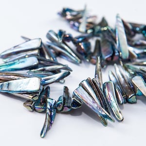 May include: Close-up of a collection of iridescent, elongated beads. The beads have a pearlescent sheen with colors ranging from blue and green to purple and silver. They are arranged on a white surface, suggesting jewelry components.