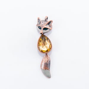 May include: A fox-shaped pendant with a silver and copper finish. The fox's head is silver with green eyes, and a large, teardrop-shaped, amber-colored gem forms the body. The tail is copper and gray. A unique piece of jewelry.