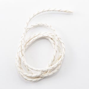 Może przedstawiać: Zwinięty, kremowy pleciony sznurek z subtelną metaliczną srebrną nitką. Sznurek wykonany jest z miękkiego, elastycznego materiału i nadaje się do różnych projektów rzemieślniczych i jubilerskich.
