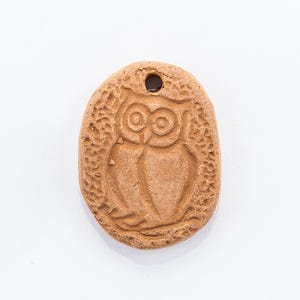 Può includere: Un ciondolo a forma di gufo in argilla marrone con un foro per appenderlo. Il gufo è raffigurato in un design semplice e stilizzato.