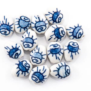 May include: A collection of white ceramic beads with a blue sunburst design. Each bead has a unique shape and is hand-painted with a detailed blue pattern. The beads are scattered on a white background.