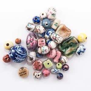 May include: A collection of ceramic beads in various shapes and sizes. The beads feature hand-painted floral, geometric, and abstract designs in shades of blue, pink, green, and brown. Some beads have text. Ideal for jewelry making or crafts.