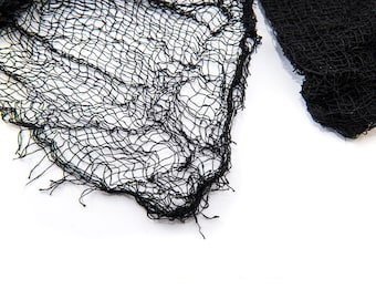 May include: Black, open-weave fabric with a distressed, frayed edge. The material has a net-like appearance and is likely used for Halloween or other themed decorations. The fabric is set against a white background.