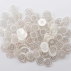May include: A collection of square, pearlescent buttons with four holes each. The buttons are in varying shades of white and gray, scattered on a white background. These buttons are ideal for sewing and craft projects.
